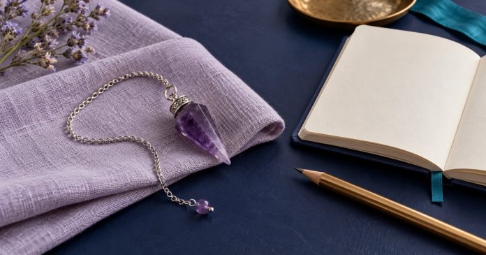 Purple crystal pendulum and silver chain resting on lavender linen beside an open notebook and gold-colored pencil