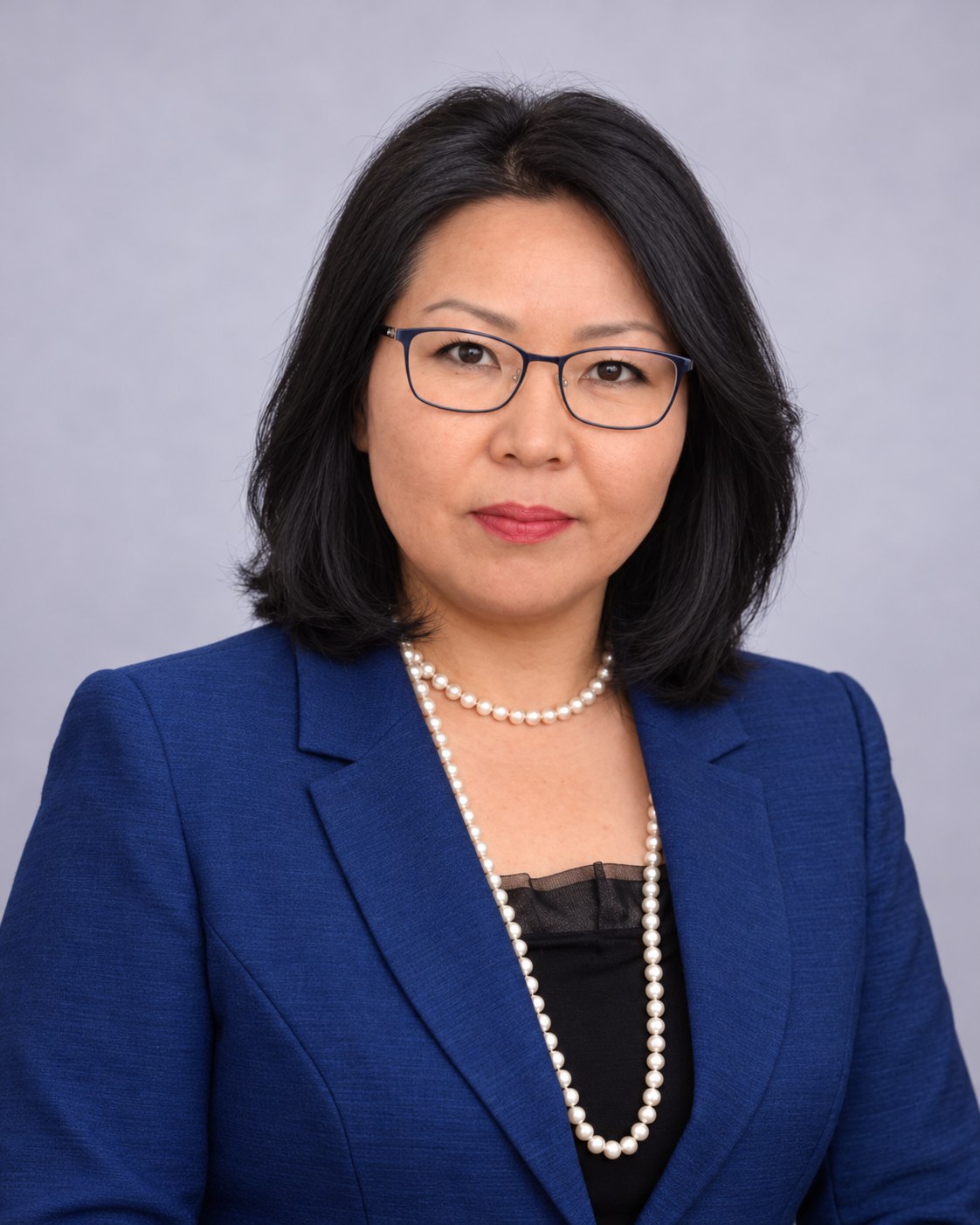 Woman wearing navy glasses, a blue jacket and pearl necklaces against a soft lavender background.